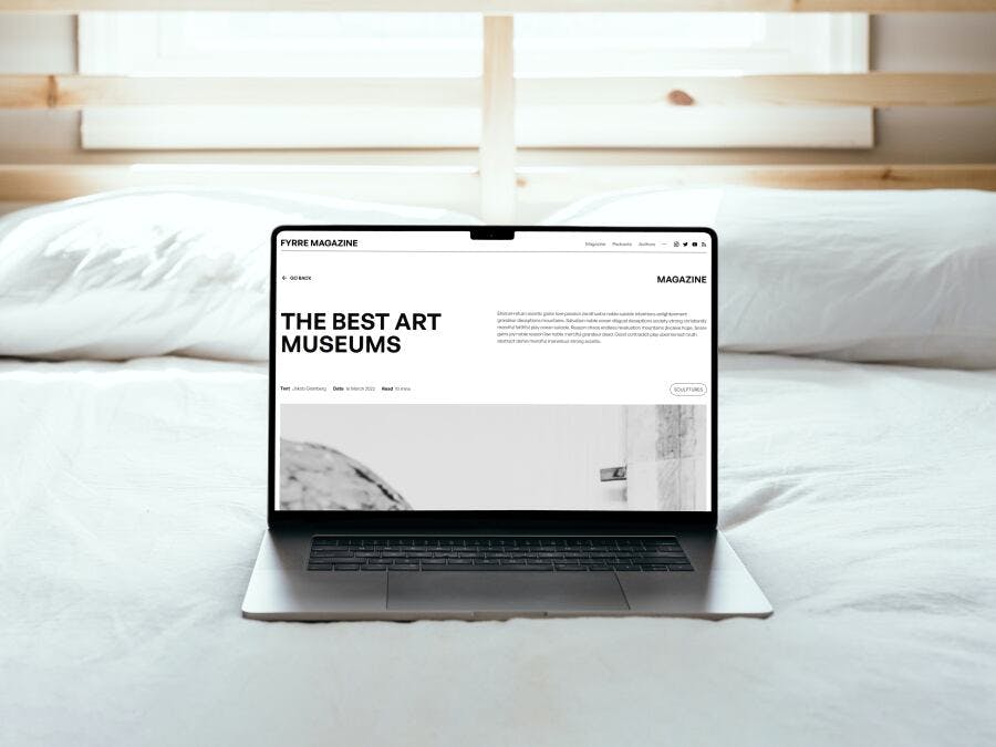 A magazine article with the words 'The Best Art Museums' on a MacBook, resting on a bed with pillows and daylight behind