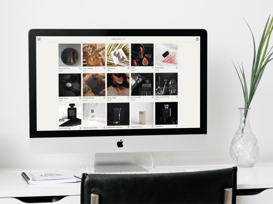 A luxury e-commerce site on an iMac, sitting on a desk with a vase to the right and a journal with a pen on the left