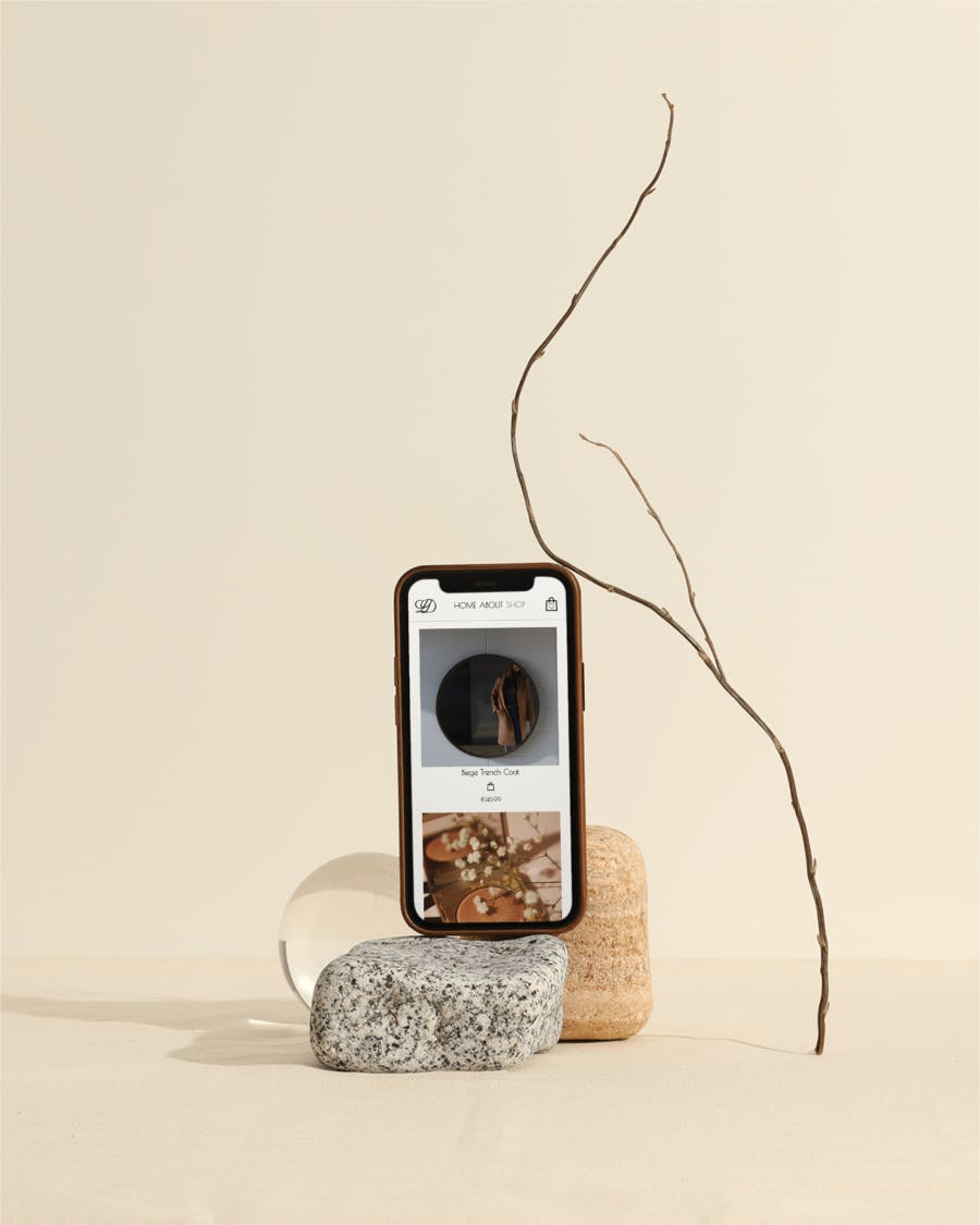 A luxury e-commerce site on a mobile device, standing upright on top of rocks, with a branch next to it and a biege backdrop